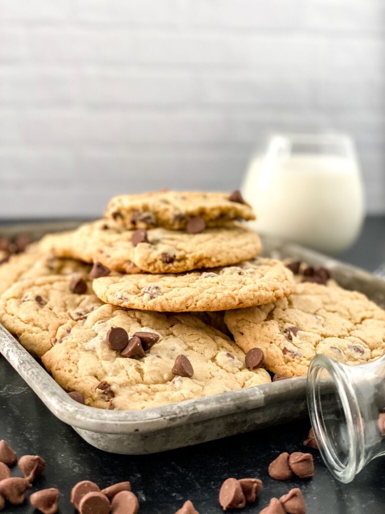 cookies stacked on a metal tray with spilled chocolate chips in the front and a cup of milk behind